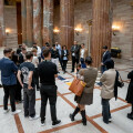 A group of people stands in a circle inside a grand hall with marble columns. They appear to be engaged in a discussion or presentation as they look at a display on the floor. The hall features ornate woodwork and tall, decorative pillars.