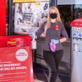 A store employee wearing a black mask and gloves stands outside a Penny Markt entrance. She holds a spray bottle and cloth next to a red sign detailing hygiene measures. An automatic sanitizer dispenser is mounted on the right.