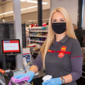 A supermarket cashier with long blonde hair is cleaning a checkout counter with a pink cloth and disinfectant spray. She is wearing a black face mask, a gray uniform with red accents, and blue gloves. A sign behind her reads "Kassa in Pause."