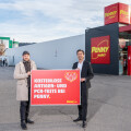 Two men outside a Penny Markt store holding a sign that reads "Kostenlose Antigen- und PCR-Tests bei Penny," promoting free testing services. The store has red signage, and the sky is partly cloudy.