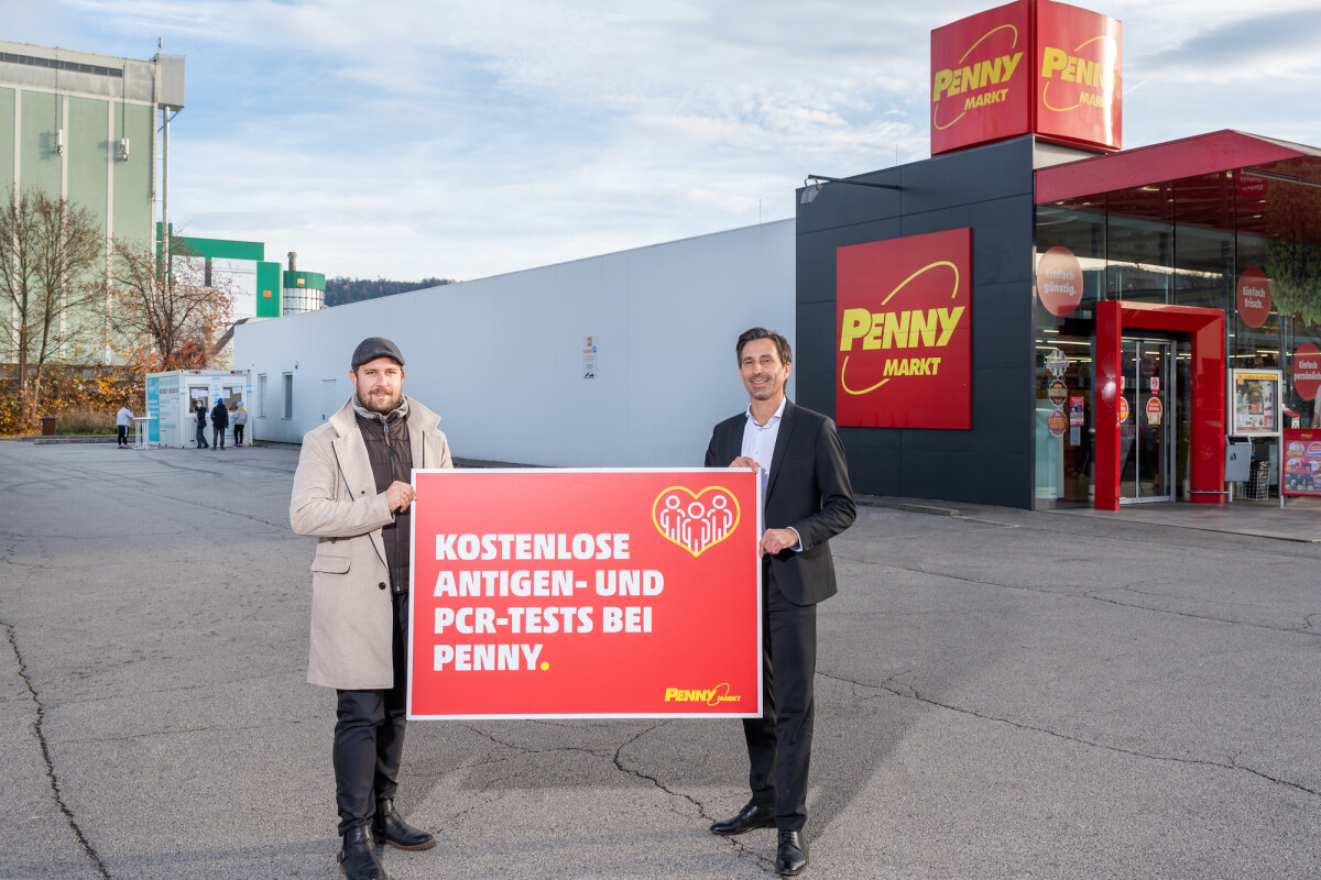 Two men outside a Penny Markt store holding a sign that reads "Kostenlose Antigen- und PCR-Tests bei Penny," promoting free testing services. The store has red signage, and the sky is partly cloudy.