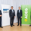 Four men in suits stand smiling in front of two vertical banners for OMV and ADEG. The backdrop is a white curtain.