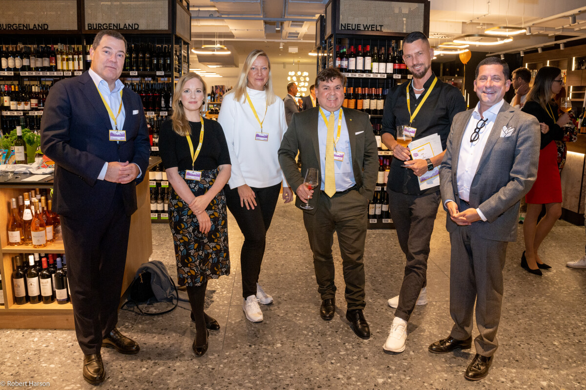 Six people stand together in a warmly lit wine store, each wearing lanyards. Bottles of wine are displayed on shelves in the background. They appear to be attending an event, with a mix of formal and casual attire.