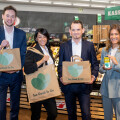 Four people smiling and holding shopping bags with the "Too Good To Go" logo in a grocery store. One person is holding a smartphone displaying an app. They are standing in front of a produce section.