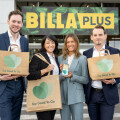 Four people in business attire stand in front of a BILLA Plus store, each holding "Too Good To Go" bags. One person holds a smartphone displaying the same app. They are smiling, and shopping carts are visible in the background.