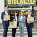 Four people stand outside a Billa Plus store holding reusable shopping bags and a smartphone showing the "Too Good to Go" app. They are dressed in business attire. The store's signs are visible above them.