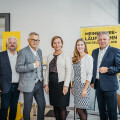 Five people standing in an office environment, all dressed in business attire. A woman and two men wear jackets, while another woman wears a white blazer over a patterned dress. They are smiling, with posters featuring yellow backgrounds visible behind them.