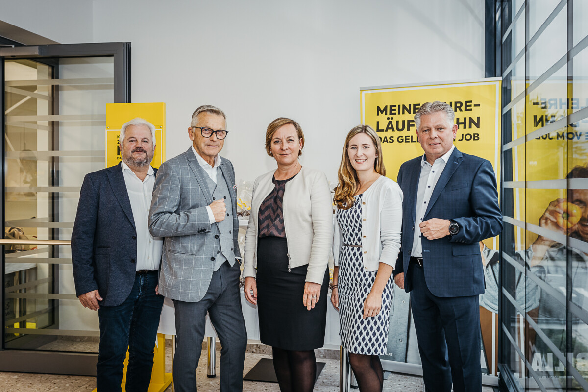 Five people standing in an office environment, all dressed in business attire. A woman and two men wear jackets, while another woman wears a white blazer over a patterned dress. They are smiling, with posters featuring yellow backgrounds visible behind them.