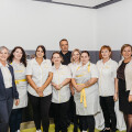 A group of people standing indoors, with several wearing matching uniforms featuring white shirts and yellow aprons, smiling towards the camera. They are posed in a well-lit room with light-colored walls.