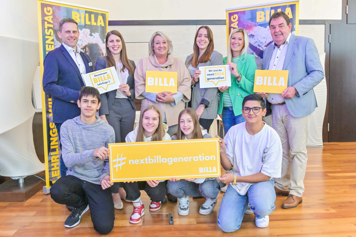 A group of people, including both adults and young adults, are holding signs with phrases like "#nextbillageneration" and "We build the next generation." They are smiling and posing together indoors, with two banners in the background.