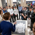 A group of people gathered around a rectangular table with a 3D architectural model, listening to a speaker. They are in a well-lit indoor space with columns, and wearing lanyards.
