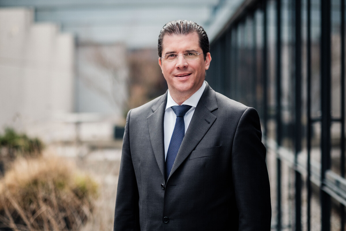 A man in a suit and tie stands outside, smiling, with a glass building in the background.