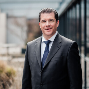 A man in a suit and tie stands outside, smiling, with a glass building in the background.