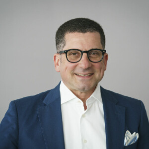A man with short hair and glasses wearing a blue suit, a white shirt, and a pocket square smiles against a plain background.