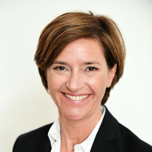 A person with short brown hair is smiling, wearing a white shirt and a black blazer against a plain background.