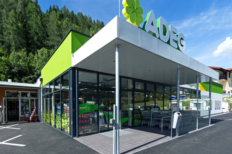 A modern supermarket with the name "ADEG" on its facade, featuring a green and white color scheme. The building is surrounded by a forested area, with shopping carts positioned near the entrance. The sky is clear with a few clouds.