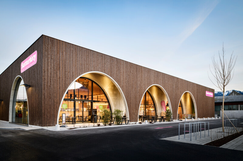 A modern, wooden commercial building with large arched windows and pink signage. The structure has a sleek, minimalist design, and there is a newly paved parking lot in the foreground. A clear blue sky is visible above.